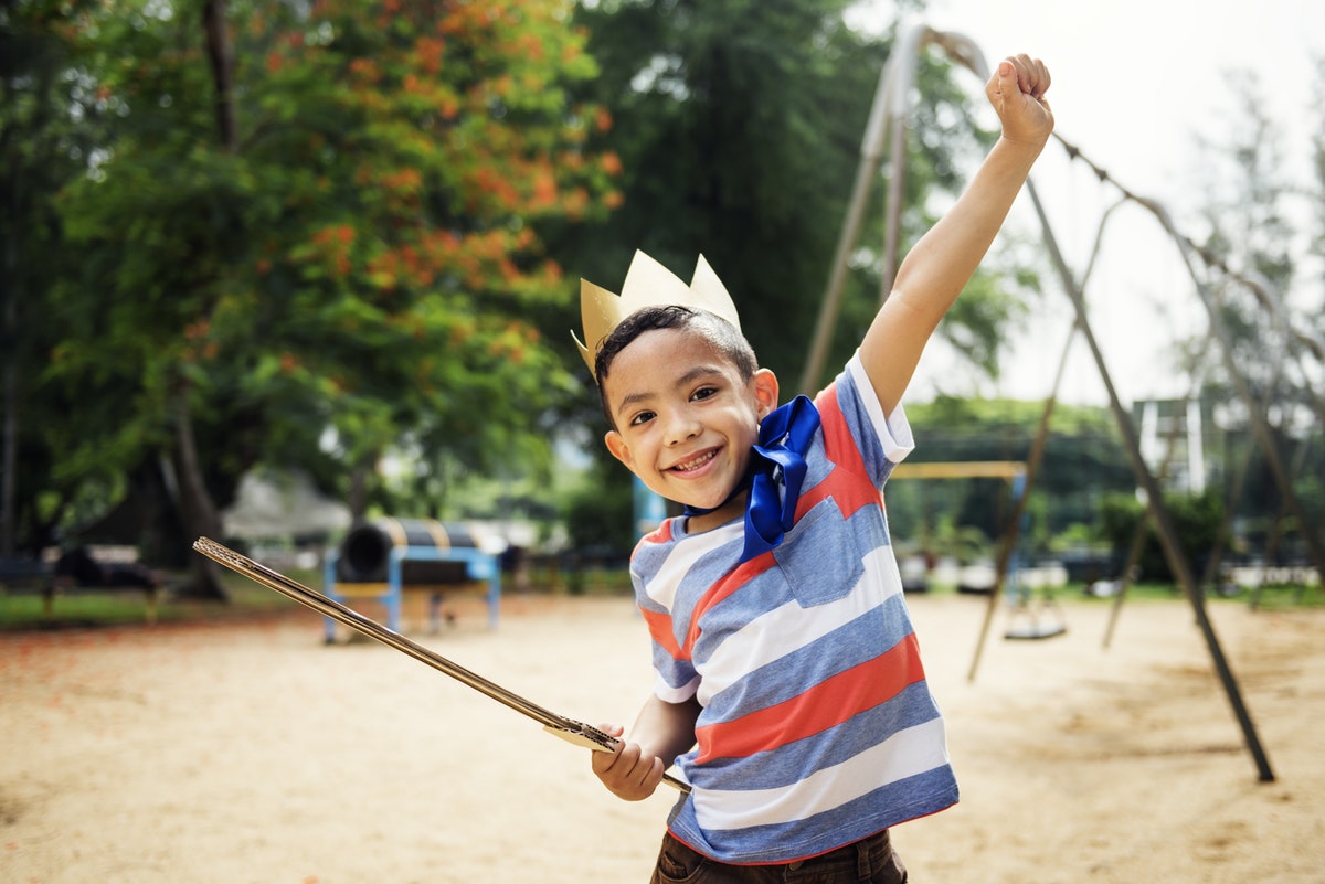 Image of a young child wearing a striped t-shirt and a paper crown, holding a toy sword with his other hand in the air.