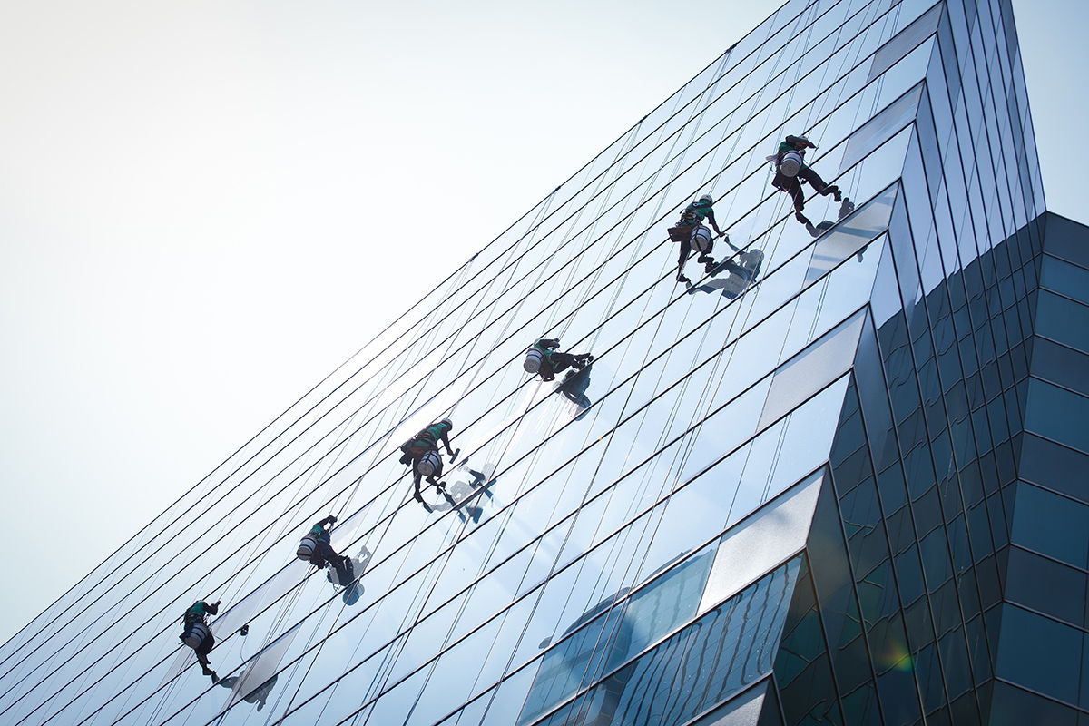 Six window cleaners with safety harnesses are suspended on the glass facade of a tall modern building, working in a row under bright sunlight. Their reflections are visible on the shiny surface.