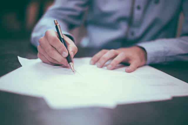 Person signing a document with a pen over scattered sheets on a desk.