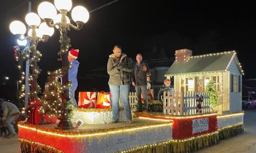 Festive parade float with a small house, lights, and gift boxes, full of people celebrating joyfully on a holiday night.