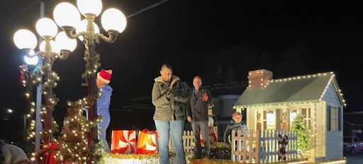 Festive parade float with a small house, lights, and gift boxes, full of people celebrating joyfully on a holiday night.