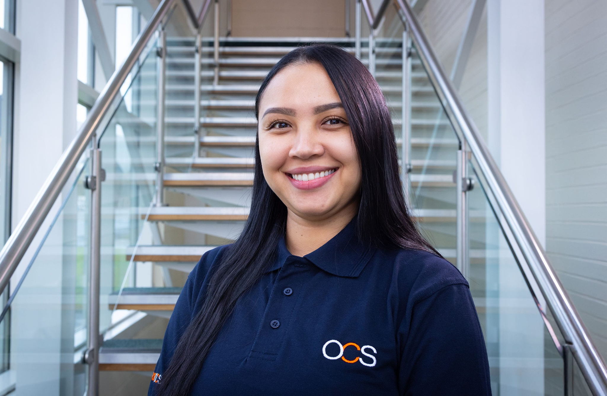 A woman with long dark hair, wearing a navy blue OCS polo shirt, stands smiling in front of a staircase with glass railings in a bright, modern building.