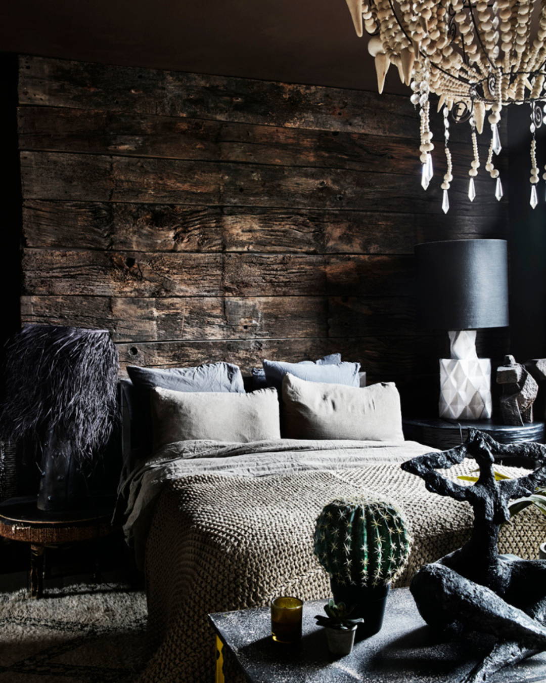 A stylish bedroom with walls made from reclaimed wood, large bedside lamps and linen bedding with a chunky wool throw.  A table at the end of the bed with plants and sculpture.