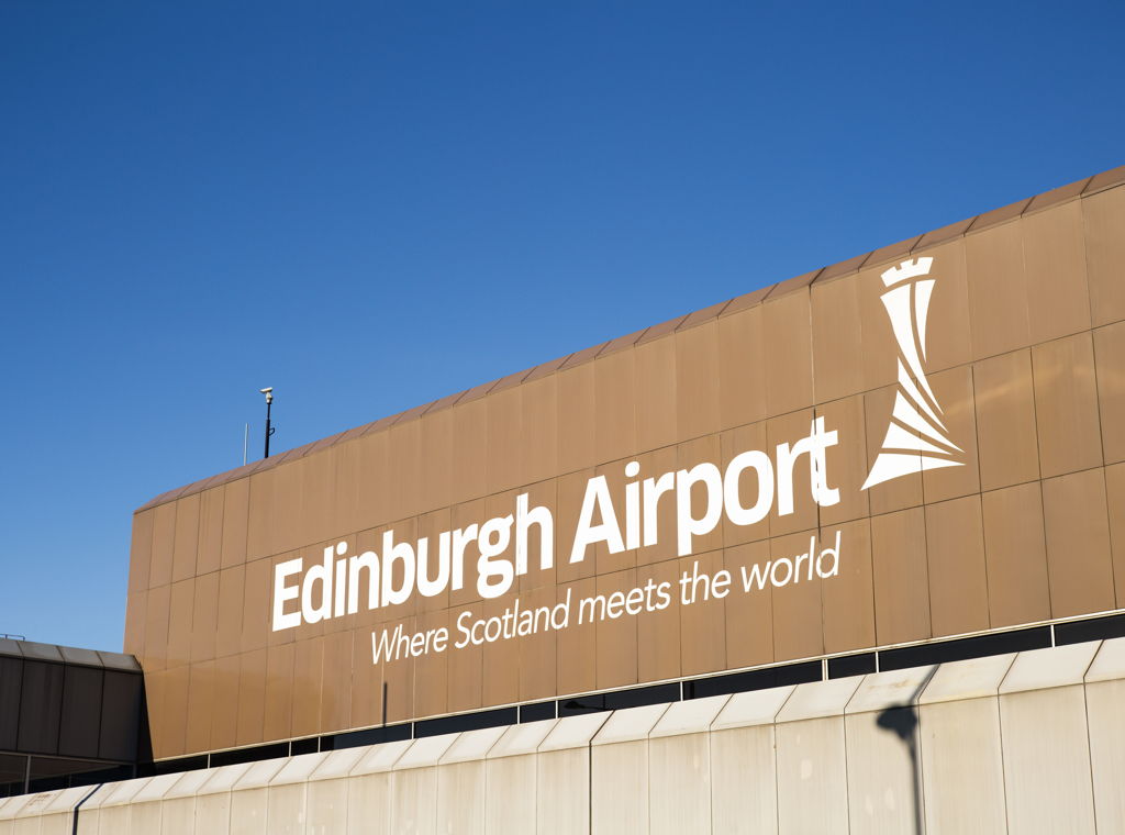Sign on the exterior of Edinburgh Airport reads Edinburgh Airport: Where Scotland meets the world, with a stylized tower logo, against a clear blue sky.