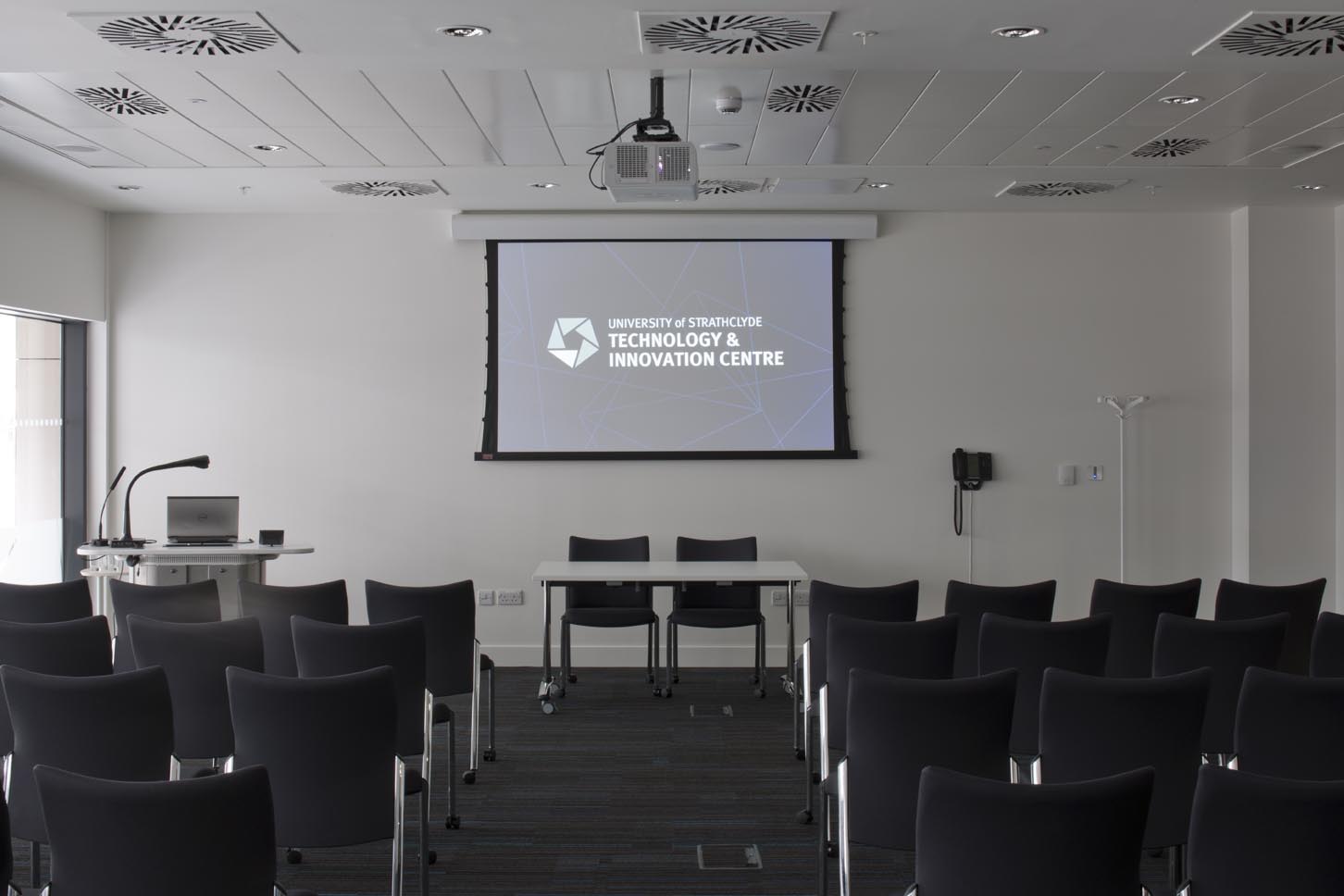 A modern conference room with black chairs facing a projector screen that reads University of Strathclyde Technology & Innovation Centre. The room has a lectern, tables, a projector, and wall-mounted equipment.