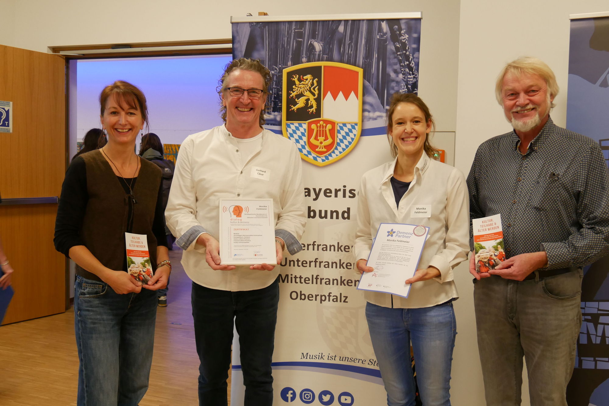 Zwei Frauen und zwei Männer halten Urkunden in den Händen und stehen vor einem Roll-up des Nordbayerischen Musikbundes. Sie lächeln stolz in die Kamera. 