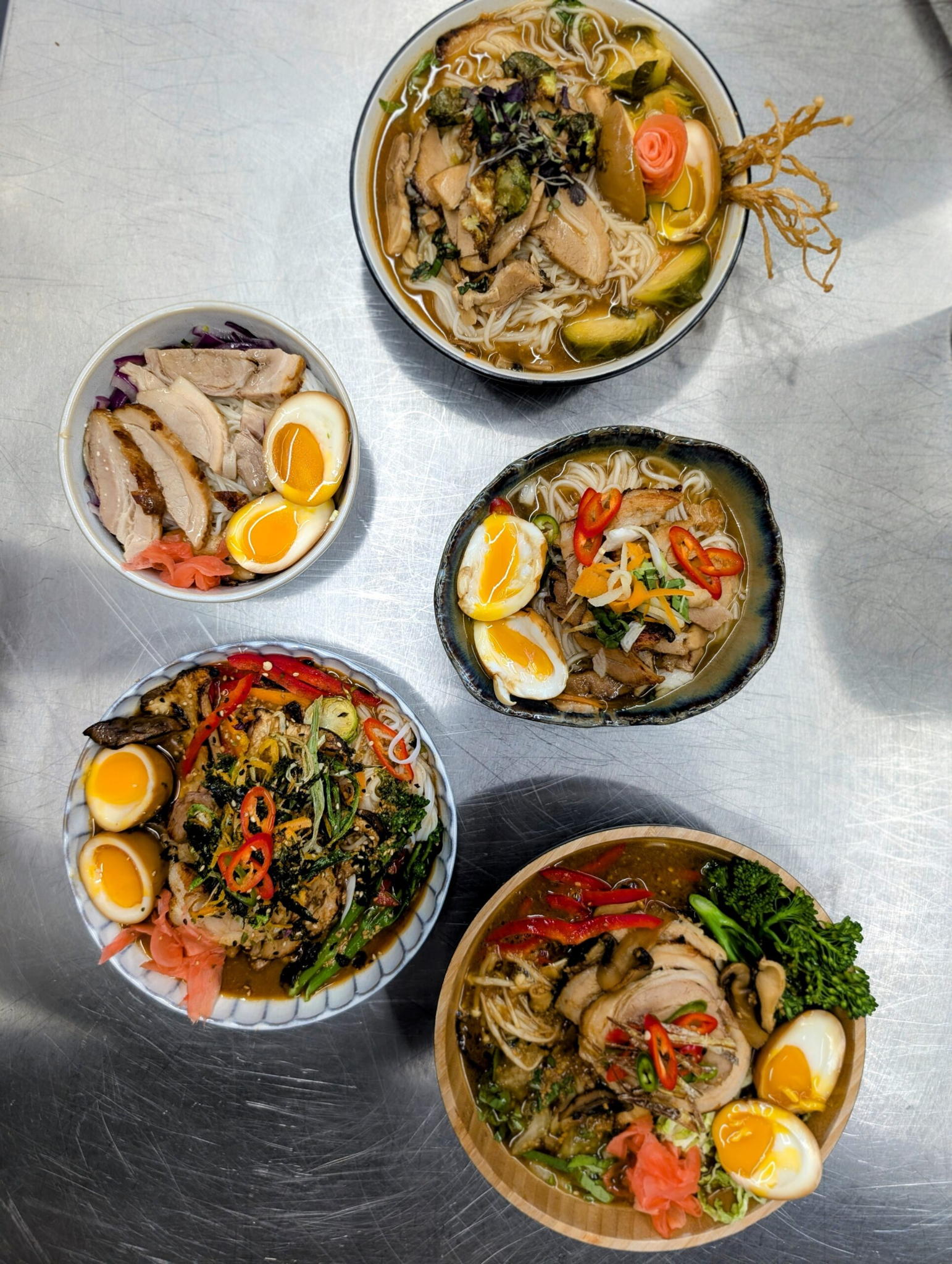 A top view of five bowls of ramen on a metal table. Each bowl is filled with noodles, sliced meat, soft-boiled eggs, and garnishes like green onions, chili peppers, and seaweed. The dishes have colorful toppings, creating a vibrant display.