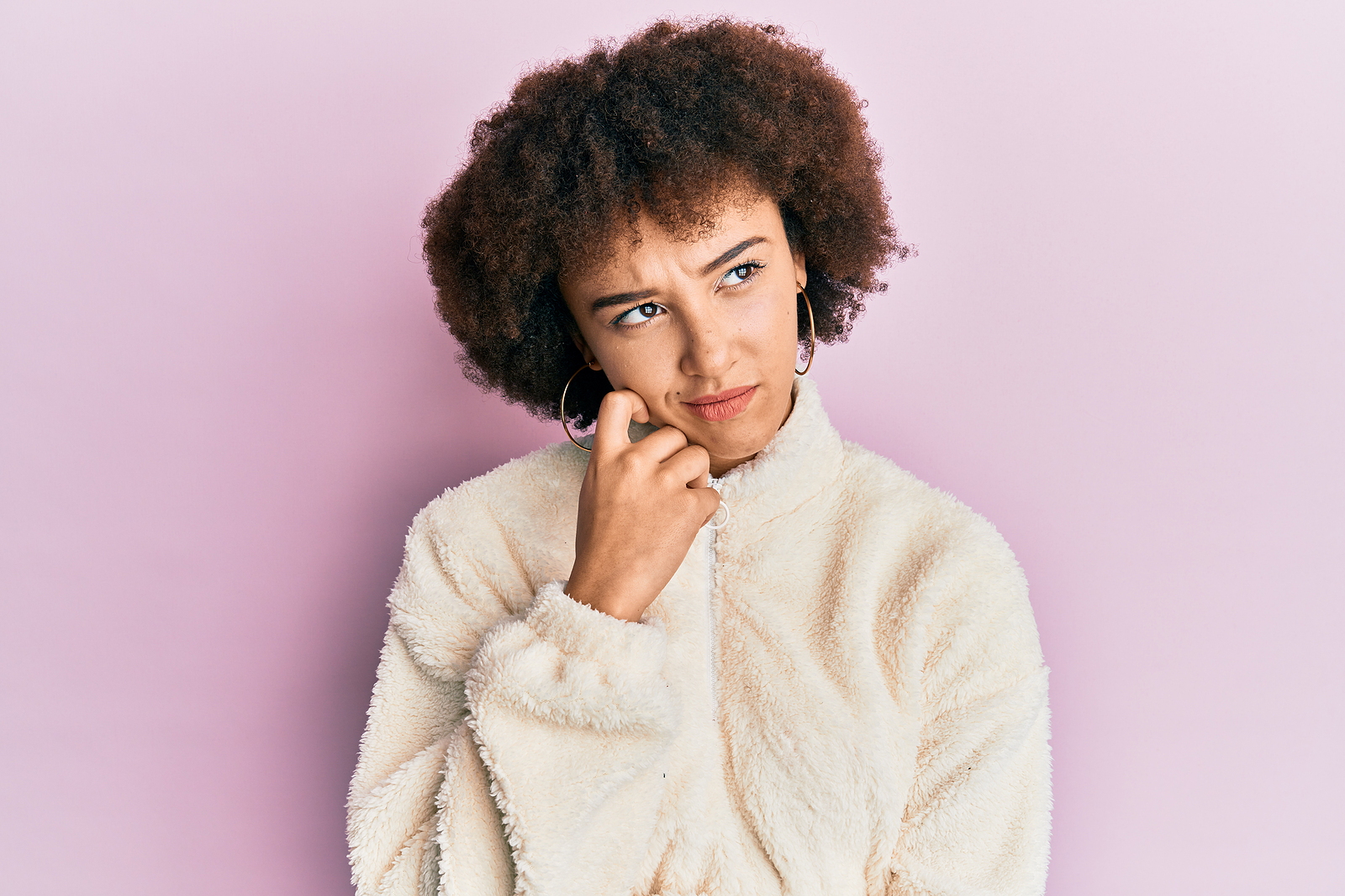 An attractive woman with large curls and puffy sweater looks pensive to the side. In front of a plain background.