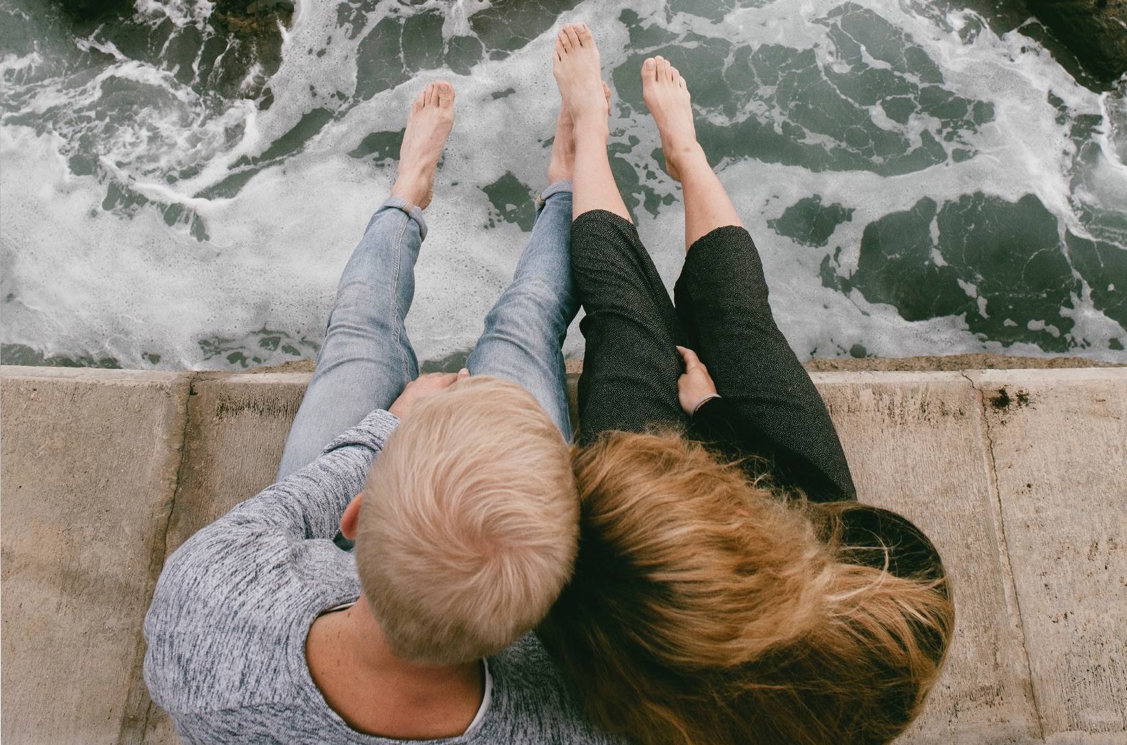 Paar sitzt auf einer Mauer am Meer und lässt Füße baumeln