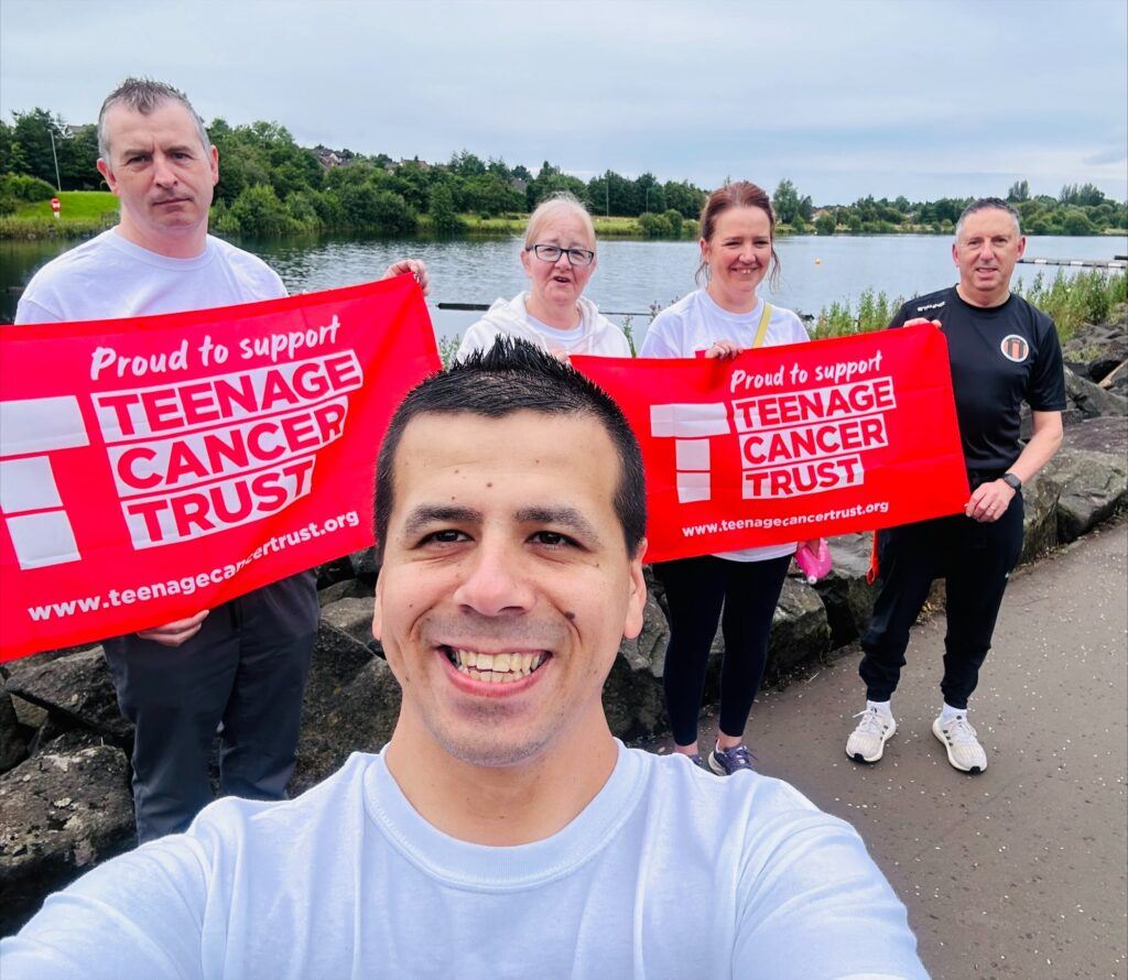 Five people stand outdoors by a lake, smiling and holding bright red banners that read Proud to support Teenage Cancer Trust. Trees and water are visible in the background.