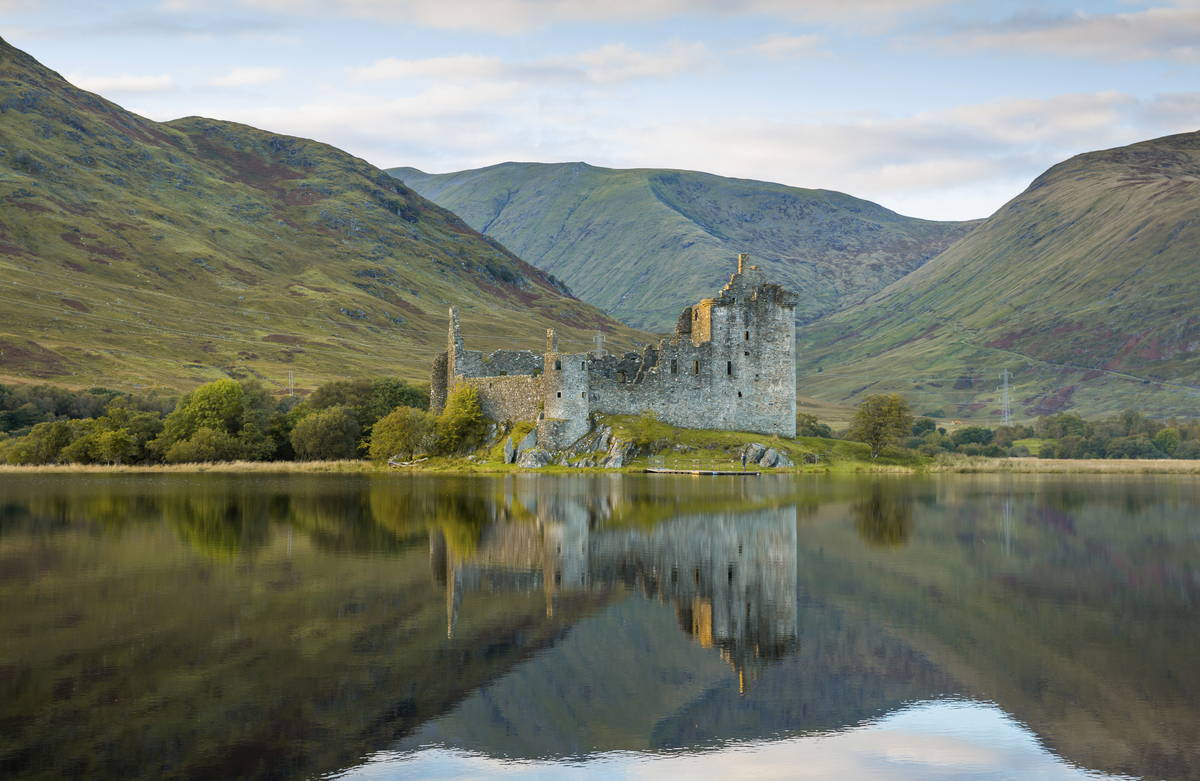 The abandoned, magnificent Kilchurn Castle, on the shore of a mirror calm Loch Awe with mountains behind and the evening sky.