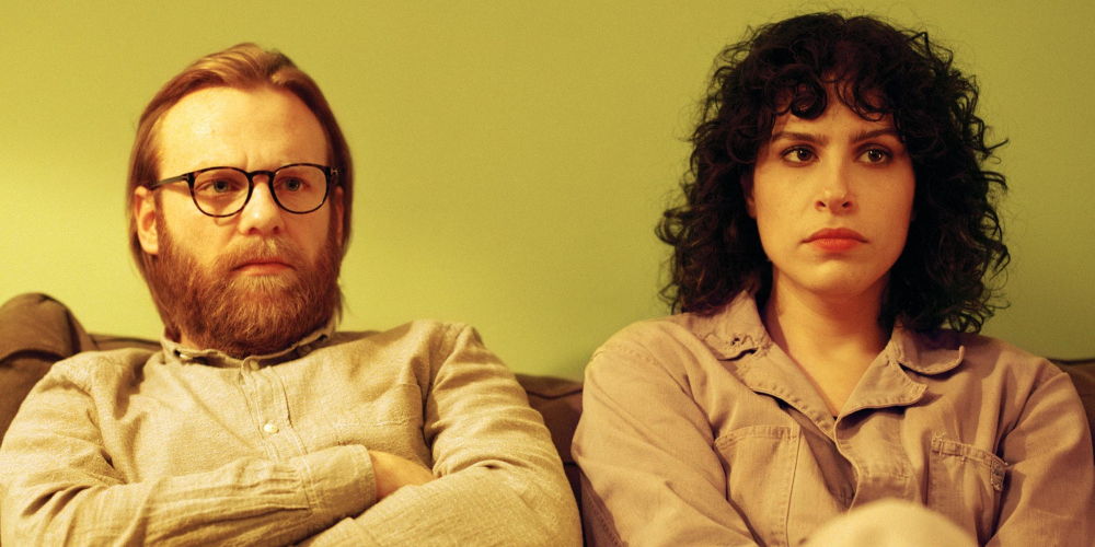 Gabe and Leila sitting on a couch both with serious expressions and arms crossed.