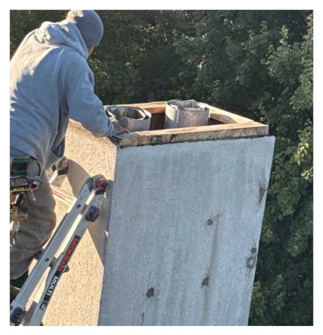Technician on a ladder inspecting and replacing a chimney chase cover on a rooftop, with tools visible and trees in the background.