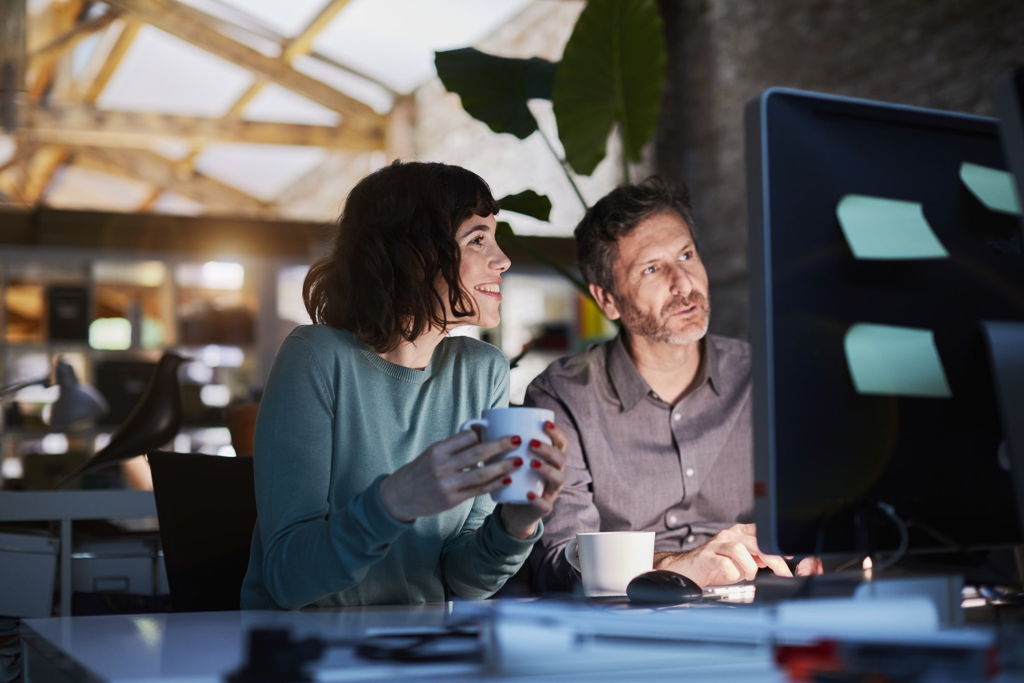 Two people sit at a desk in a modern office, looking at a computer monitor. The woman holds a mug and smiles, while the man looks focused. The office is dimly lit with natural light and large plants in the background.