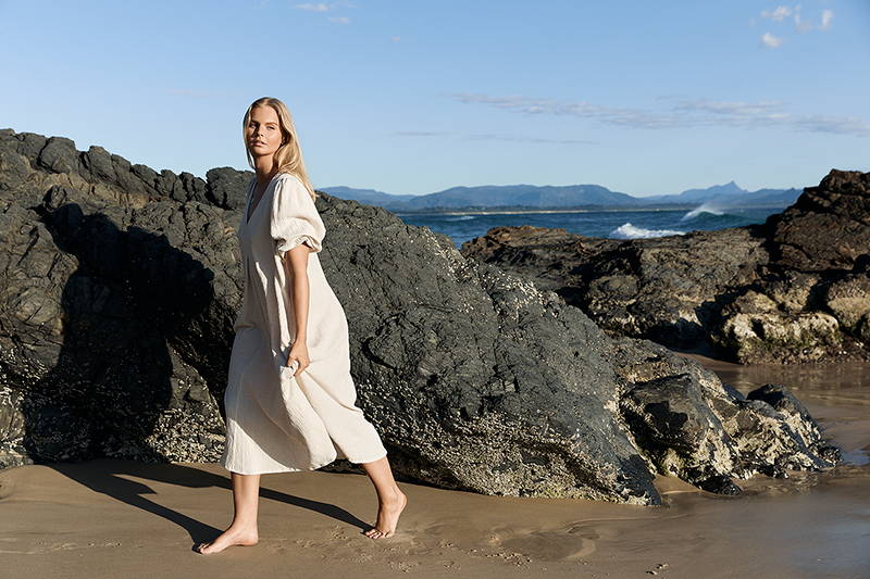 beautiful blonde woman walking barefoot on wet sand wearing loose linen dress