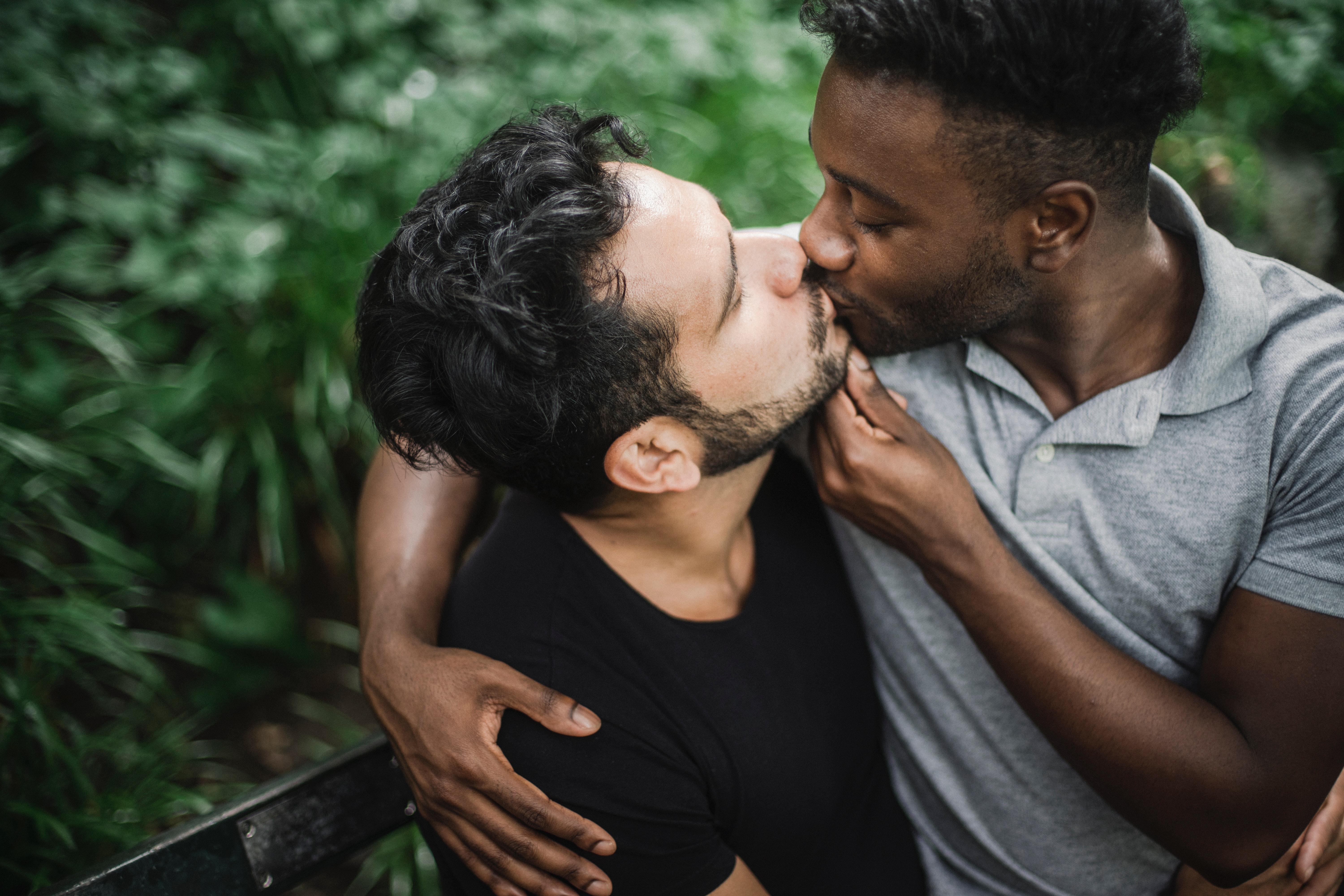 Two multi ethnic men holding eachother and kissing with a outdoor background behind them.
