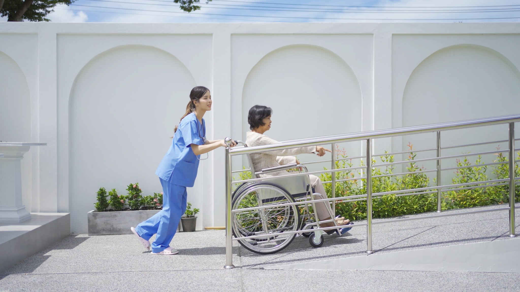 A caregiver in blue scrubs pushes an older person in a wheelchair up a ramp outdoors, beside a white wall with arched designs under a bright, sunny sky.