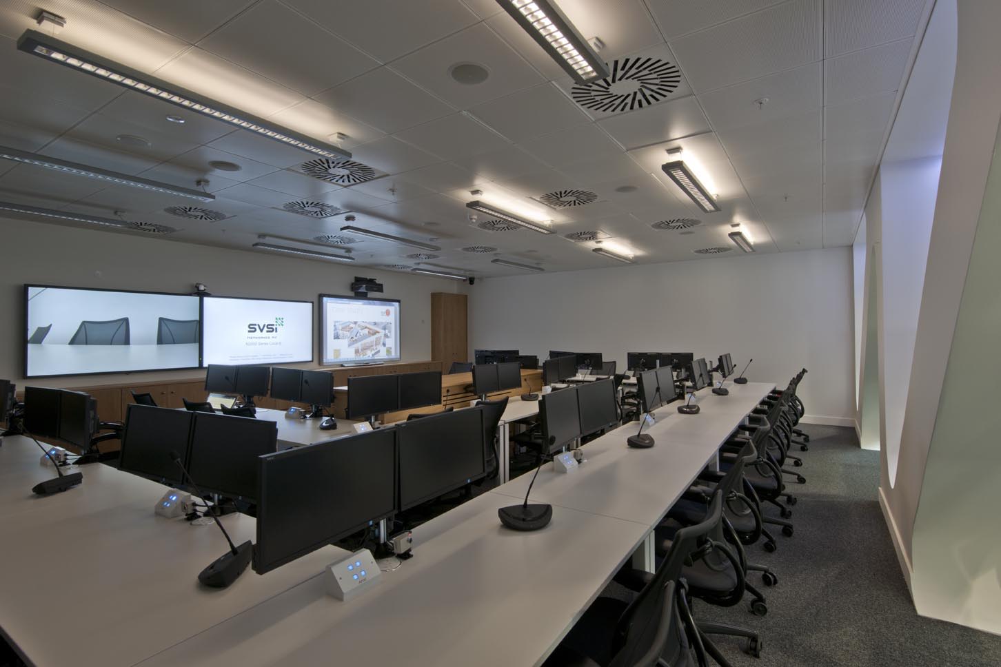 A modern conference room with rows of desks, each equipped with computer monitors, microphones, and office chairs. Two large display screens are on the front wall, and the room is brightly lit with ceiling lights.