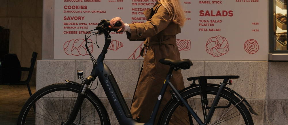a woman walking her gazelle electric city bike in front a food menu in new york city