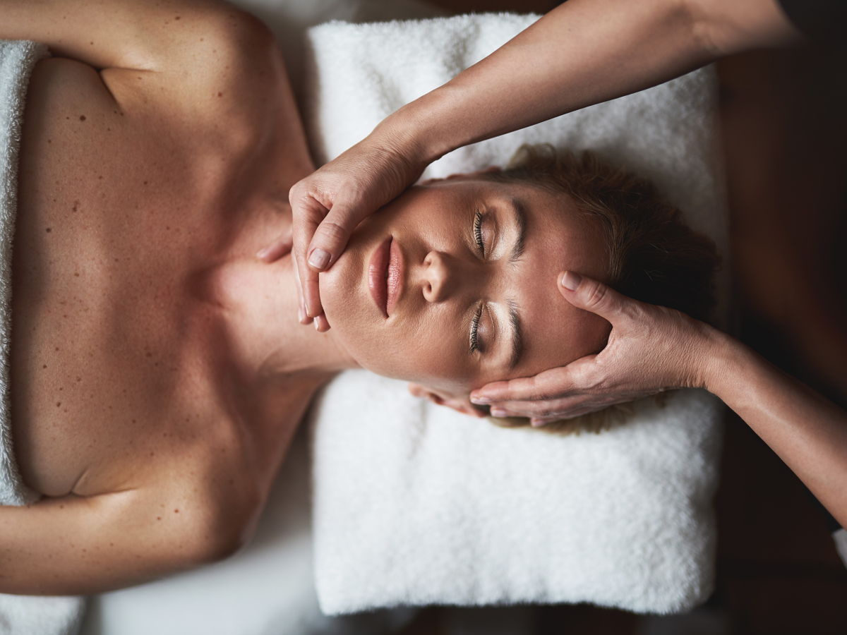 Eine entspannte Frau liegt mit dem Kopf auf einem Handtuch und bekommt eine Gesichtsmassage.