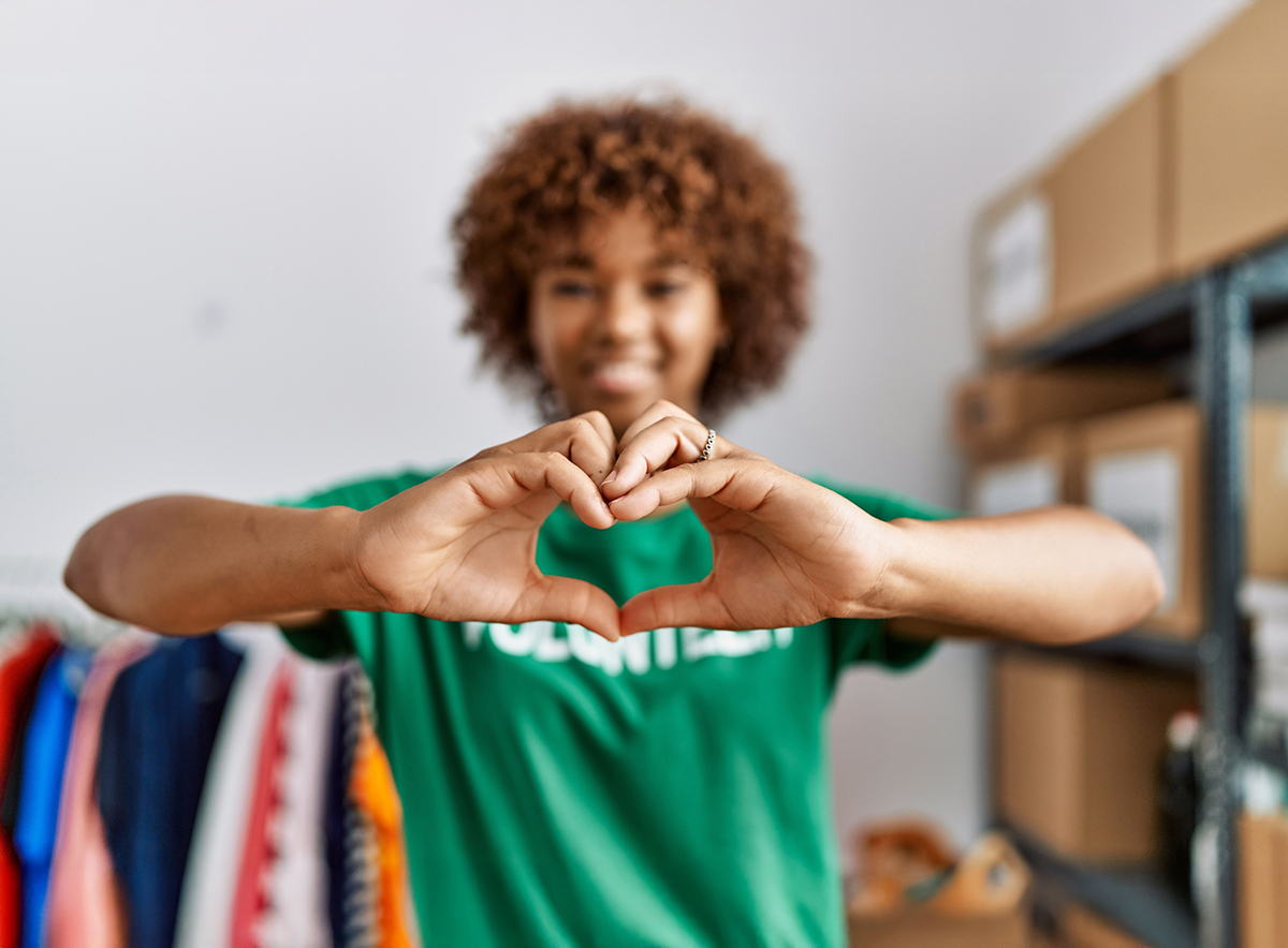 A person wearing a green Volunteer shirt forms a heart shape with their hands while standing in a room with clothing racks and shelves in the background.