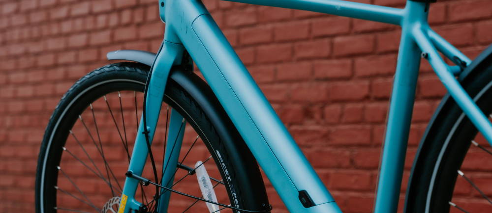 A teal Tenways electric city bike against a brick wall