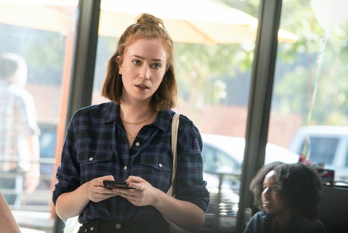 Ava wearing a flannel shirt, has her phone in her hands and is looking at someone off camera with a serious face.