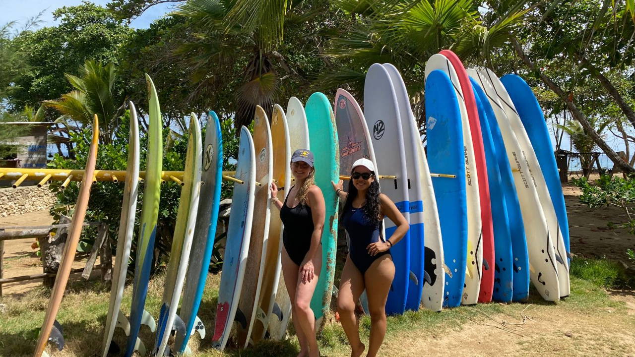 Two women in one-piece swimsuits standing with a row of surfboards on the beach in the Dominican Republic