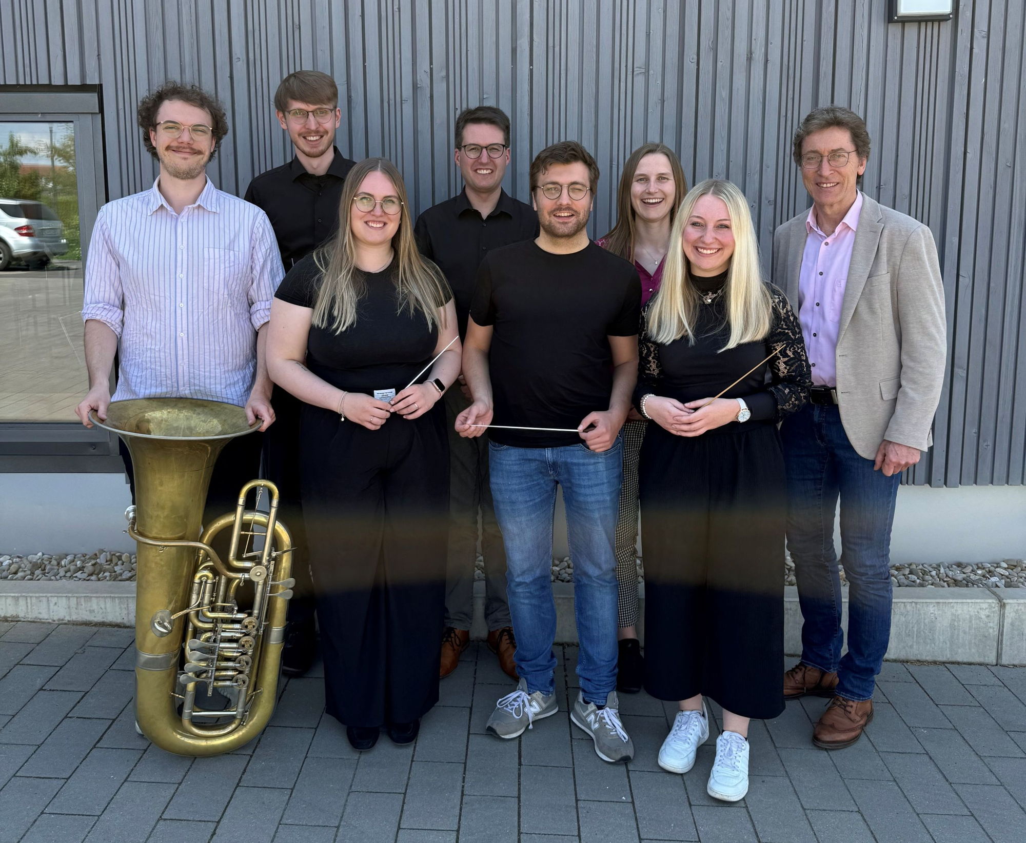 Einige Männer und Frauen stehen nebeneinander und lächeln in die Kamera. Vor einem der Männer steht eine Tuba und die anderen halten teilweise Taktstöcke in den Händen. 