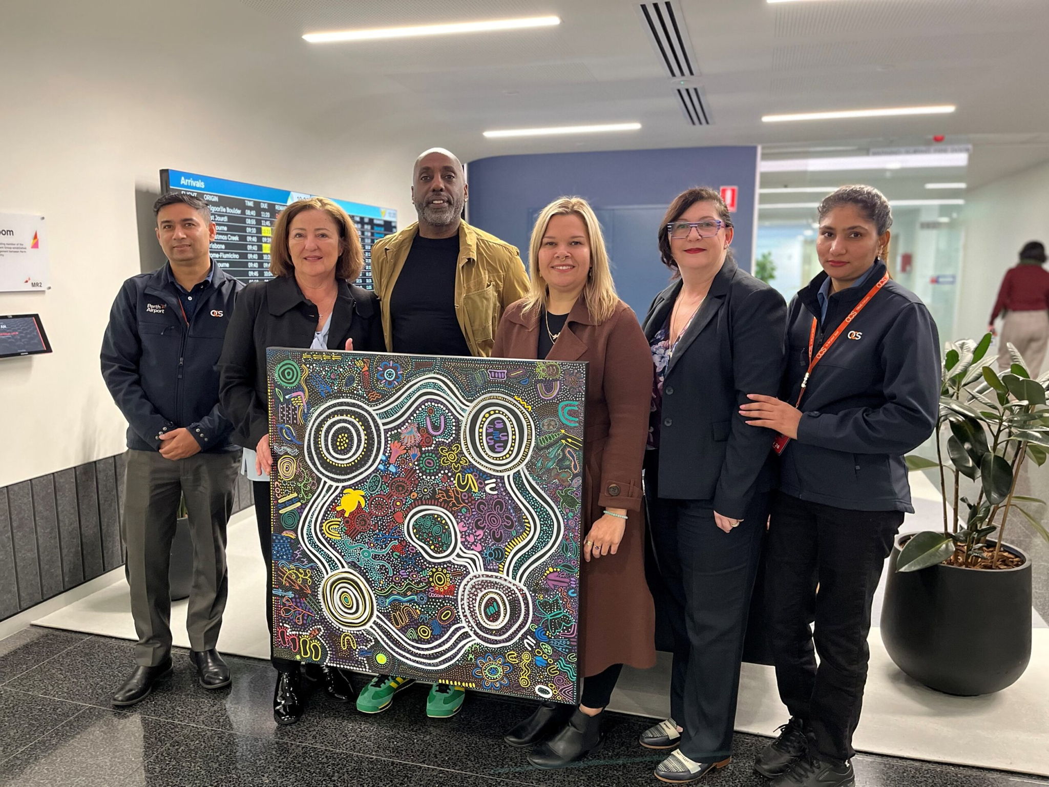 Six people stand indoors, posing together and smiling, while holding a colorful, abstract Indigenous artwork with circular patterns. They are dressed in business or business-casual attire, in a modern office hallway.