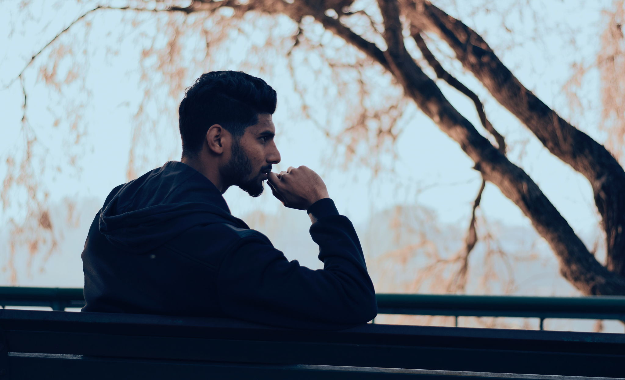 Silhouette of a man with a beard sitting on a park bench with a pensive look on his face and his hand on his chin.