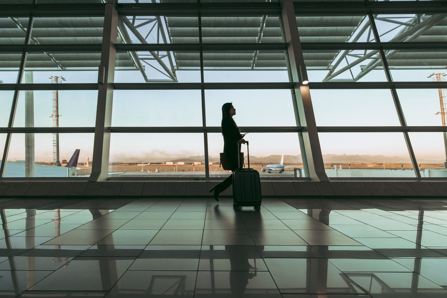 A person walks through an airport terminal, pulling a wheeled suitcase. Large windows show airplanes and a clear sky outside. The sun casts reflections on the shiny floor.