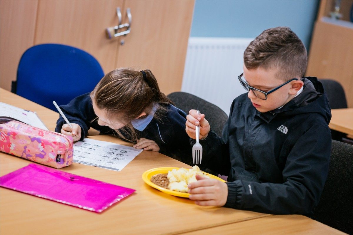 Two children sit at a table. One girl is writing on a worksheet, her head bent down. Next to her, a boy wearing glasses and a black jacket is eating food from a yellow plate with a fork. School supplies are on the table.