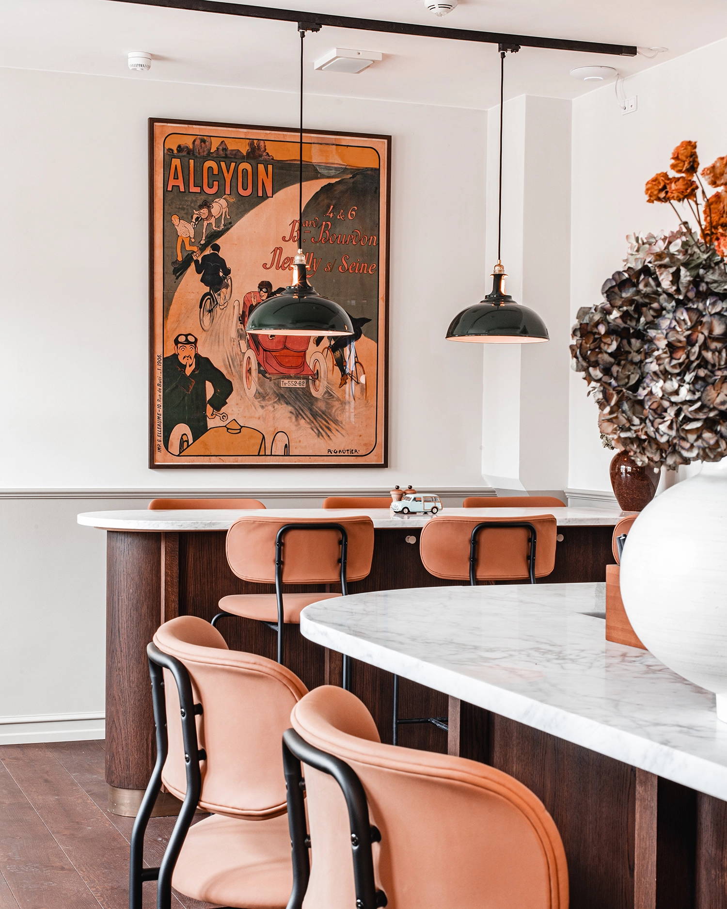 Two large Green Coolicon Pendant lampshades hanging above a bar island inside a restaurant with leather bar stools and a large framed painting on the back wall.