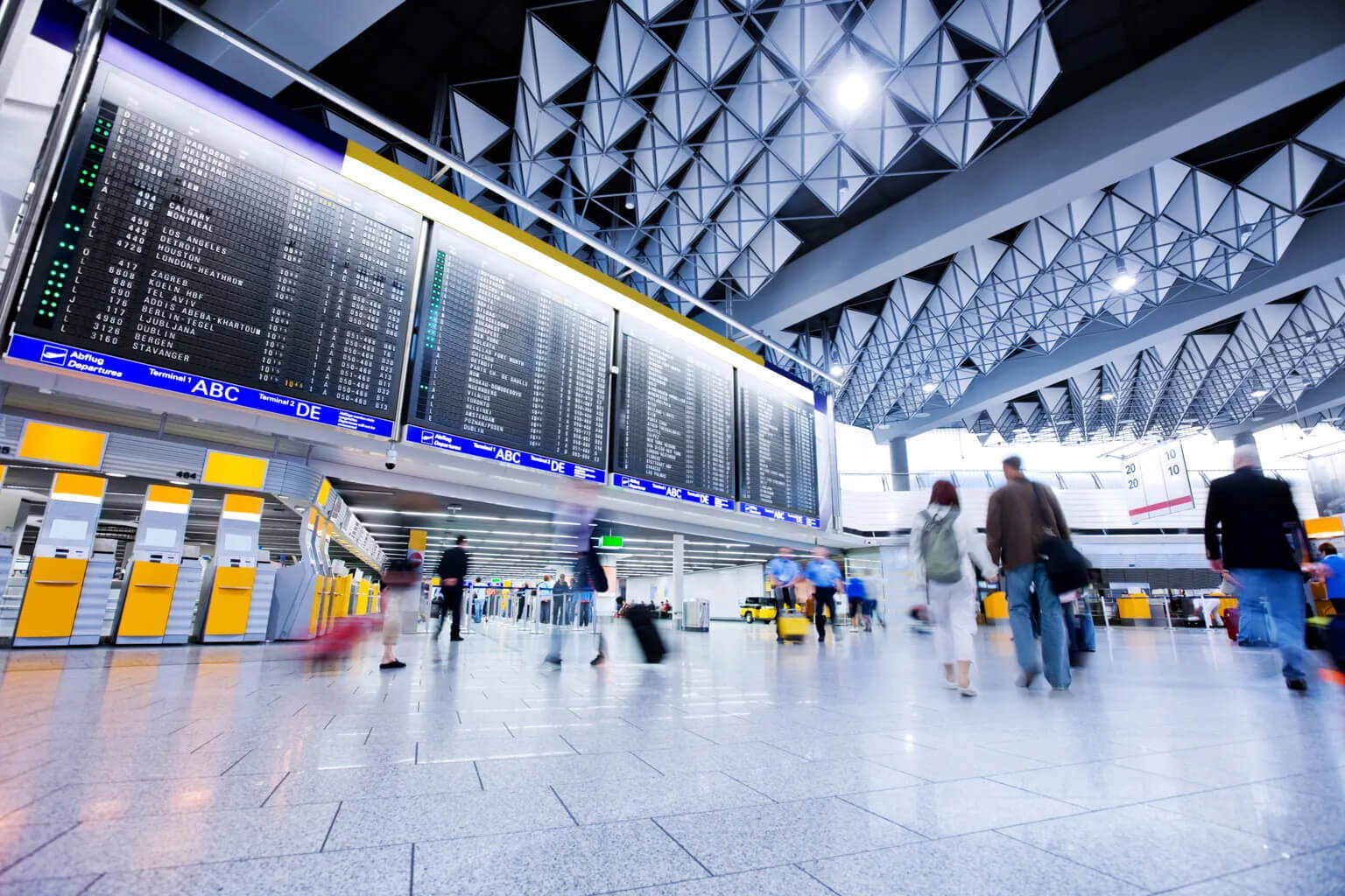 A busy airport terminal with people walking and pulling luggage, large flight information display boards overhead, and yellow check-in counters in the background.