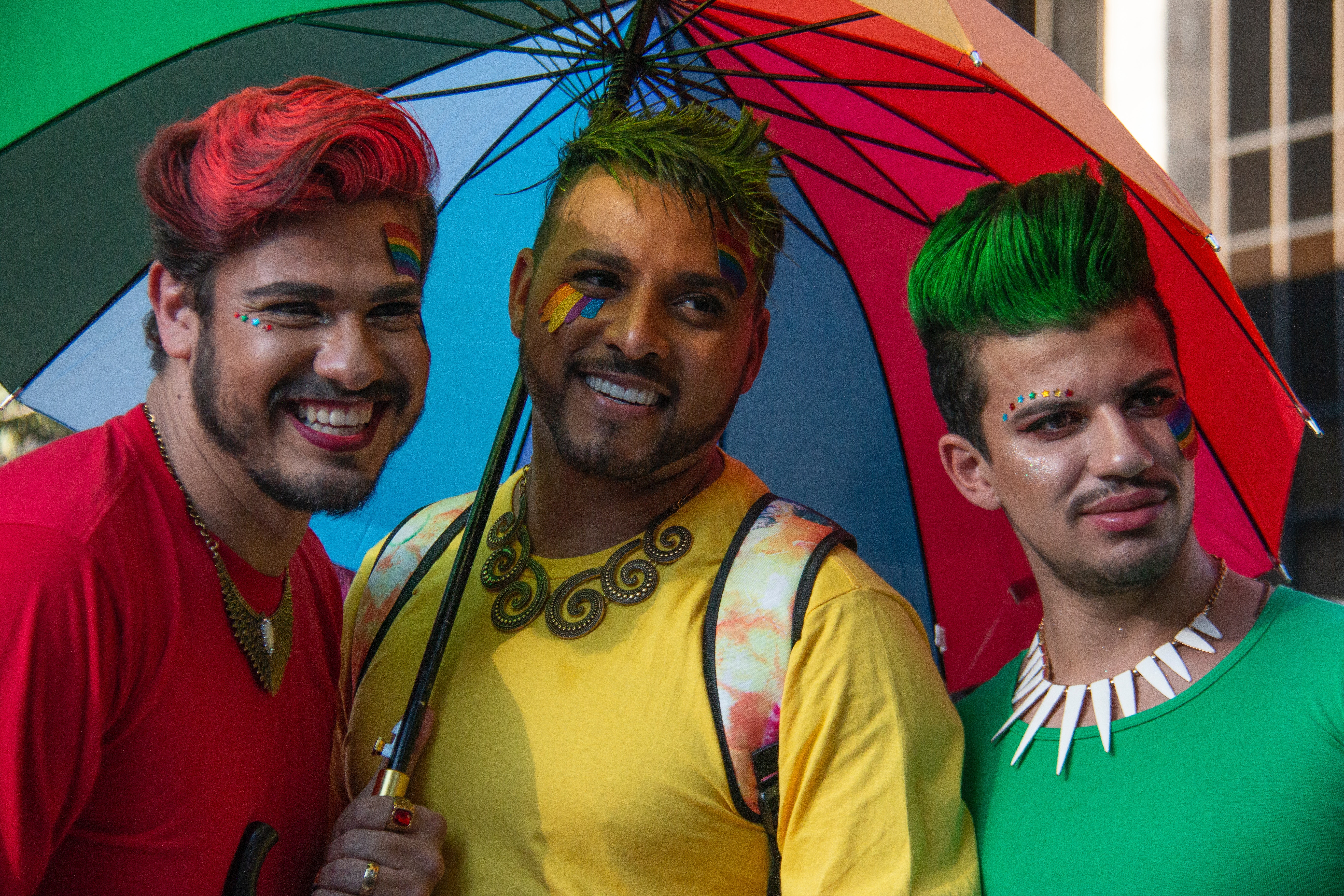 3 friends pose smiling for a picture during a pride event. They have dyed hair and colorful makeup.
