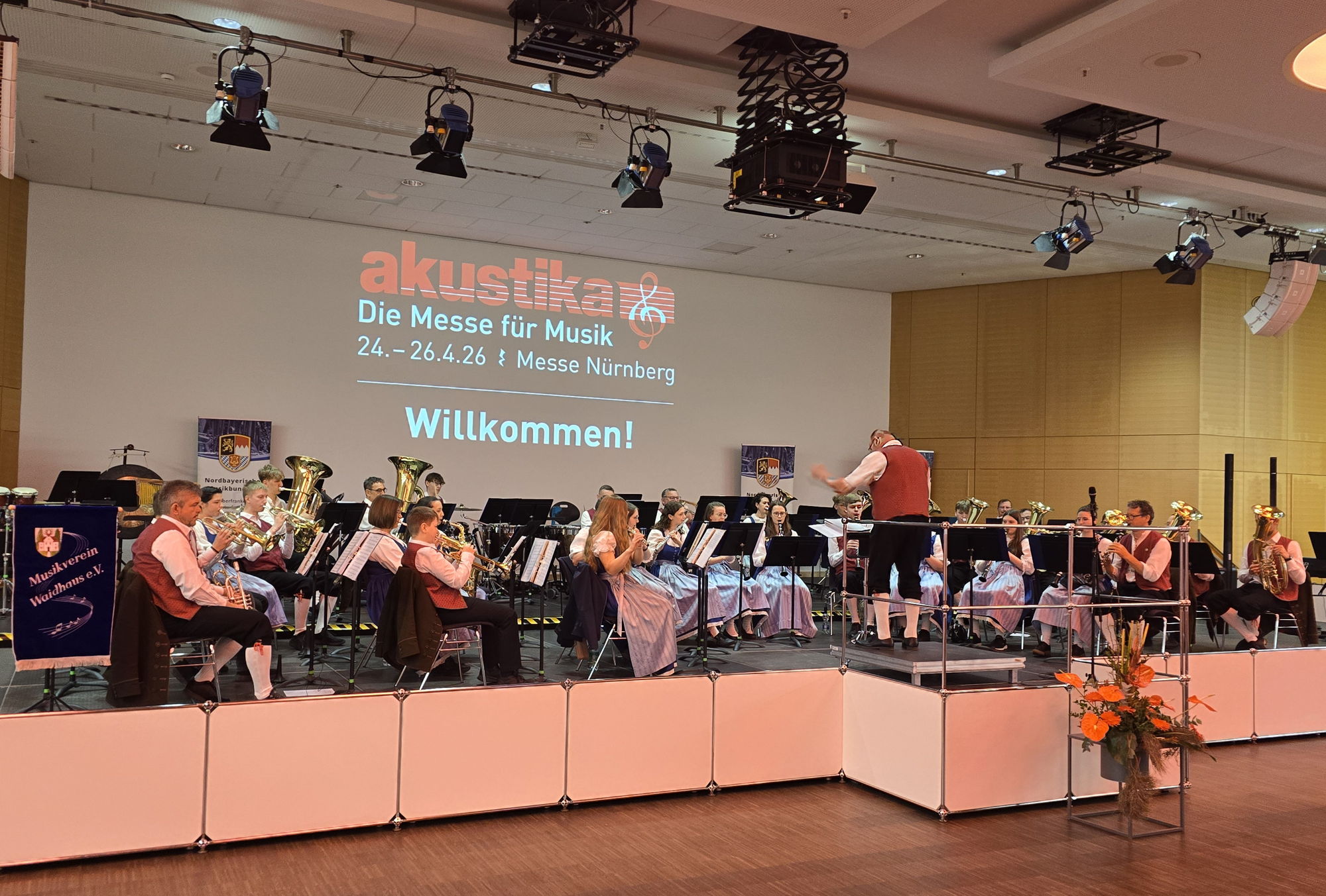 Auf einer Bühne sitzt ein Blasorchester in Tracht und musiziert. Hinter der Bühne steht eine Leinwand auf der das Logo der Musik-Messe "akustika" projiziert ist. 