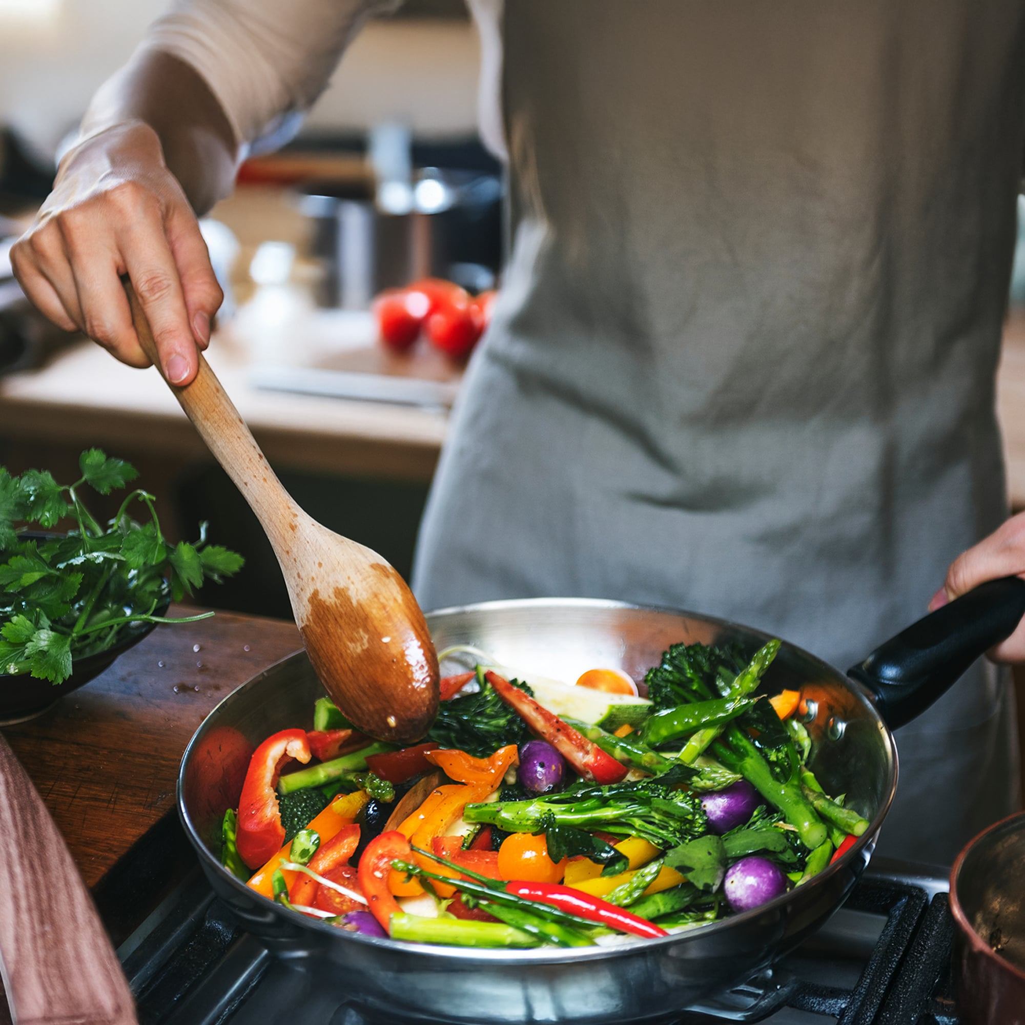Person in a gray apron stirring colorful vegetables, including bell peppers, asparagus, and cherry tomatoes, in a skillet with a wooden spoon in a kitchen setting. Fresh herbs and tomatoes are visible in the background.