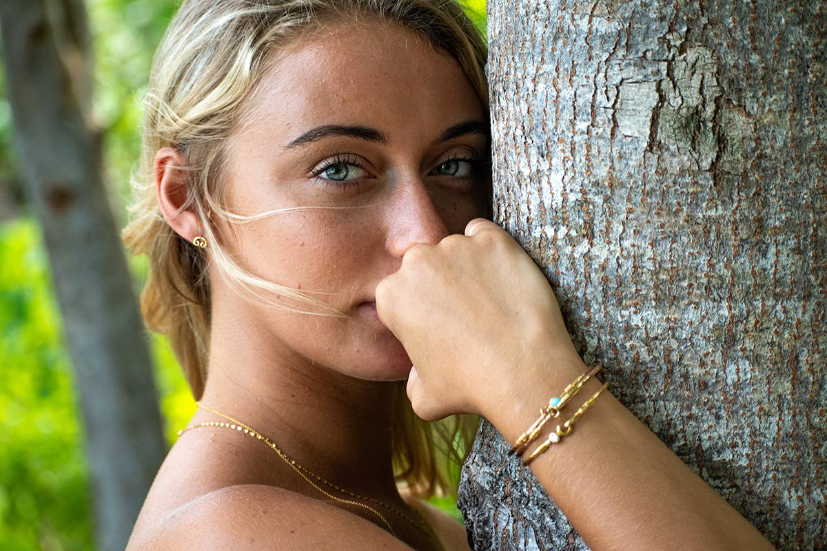 A woman standing next to a tree wearing gold jewelry.