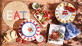 A display of summer dining alfresco featured on a wooden table in bright sunlight with some dappled light. A large straw place mat with the word EAT embroidered in white 'E', red 'A' and blue 'T', a bread board with cut bagette, two melamine plates featuring tomato illustrations on a white background, another wooden board as a platter feturing grapes, cheeses, cooked meats and cherry tomatoes, a small red rimmed bowl, a blue check napkin and a book titled Charcuterie boards.