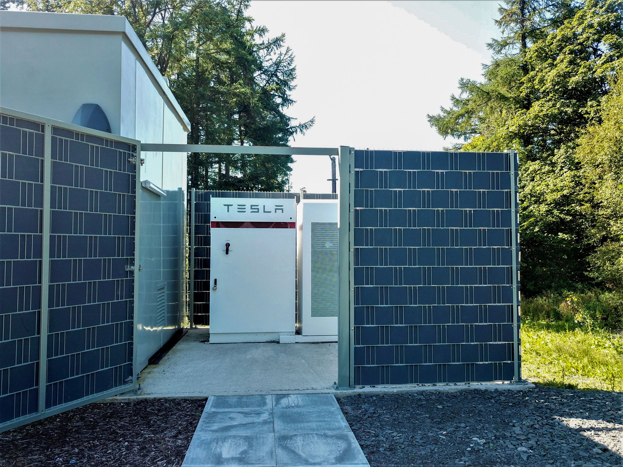 A white Tesla energy storage unit is enclosed by tall solar panel fences, with trees and greenery visible in the background under clear daylight.