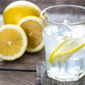 A glass of water with ice-cubes and lemon slices, with 2 lemons situated on the left hand side, one is halved and the other is whole