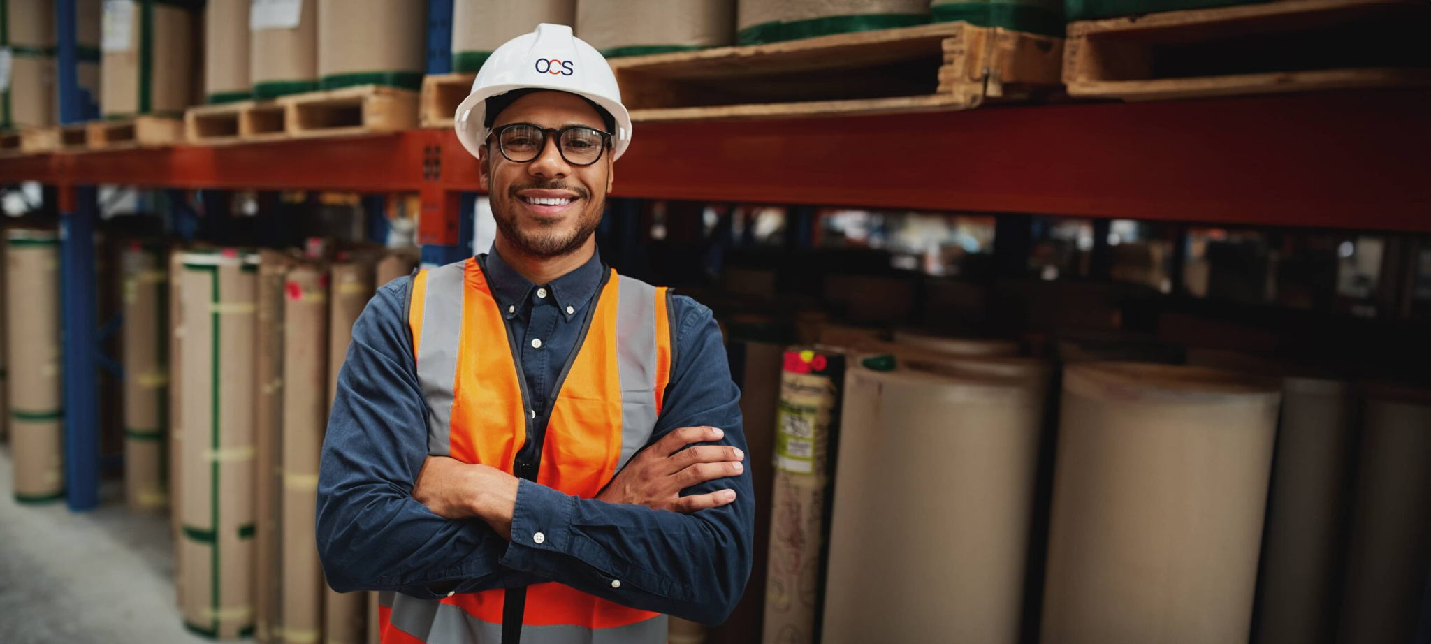 A smiling OCS colleague stands in a warehouse surrounded by tall stacks of paper,