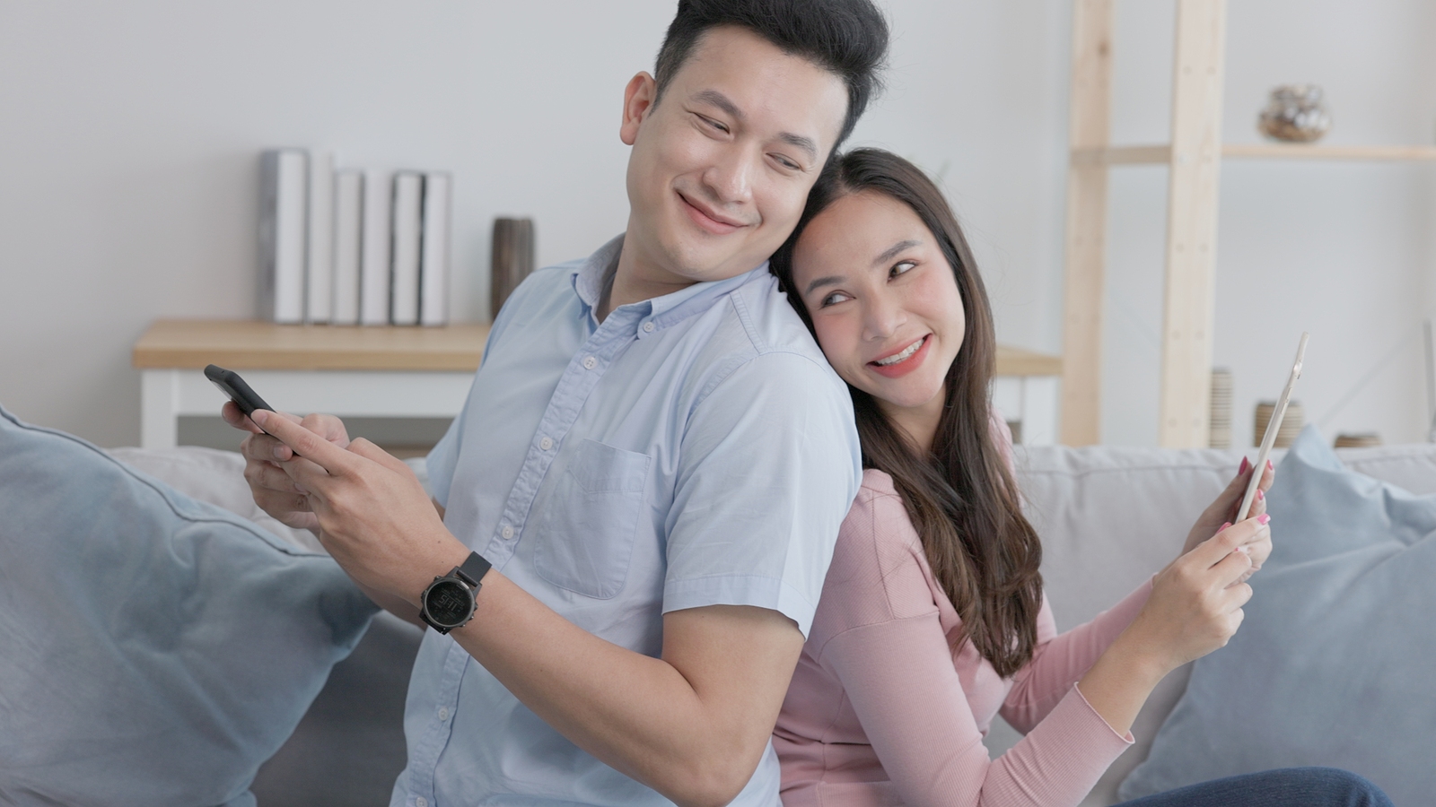 An attractive asian couple sits on the couch back to back smiling looking over eachothers shoulders holding each a cellphone.