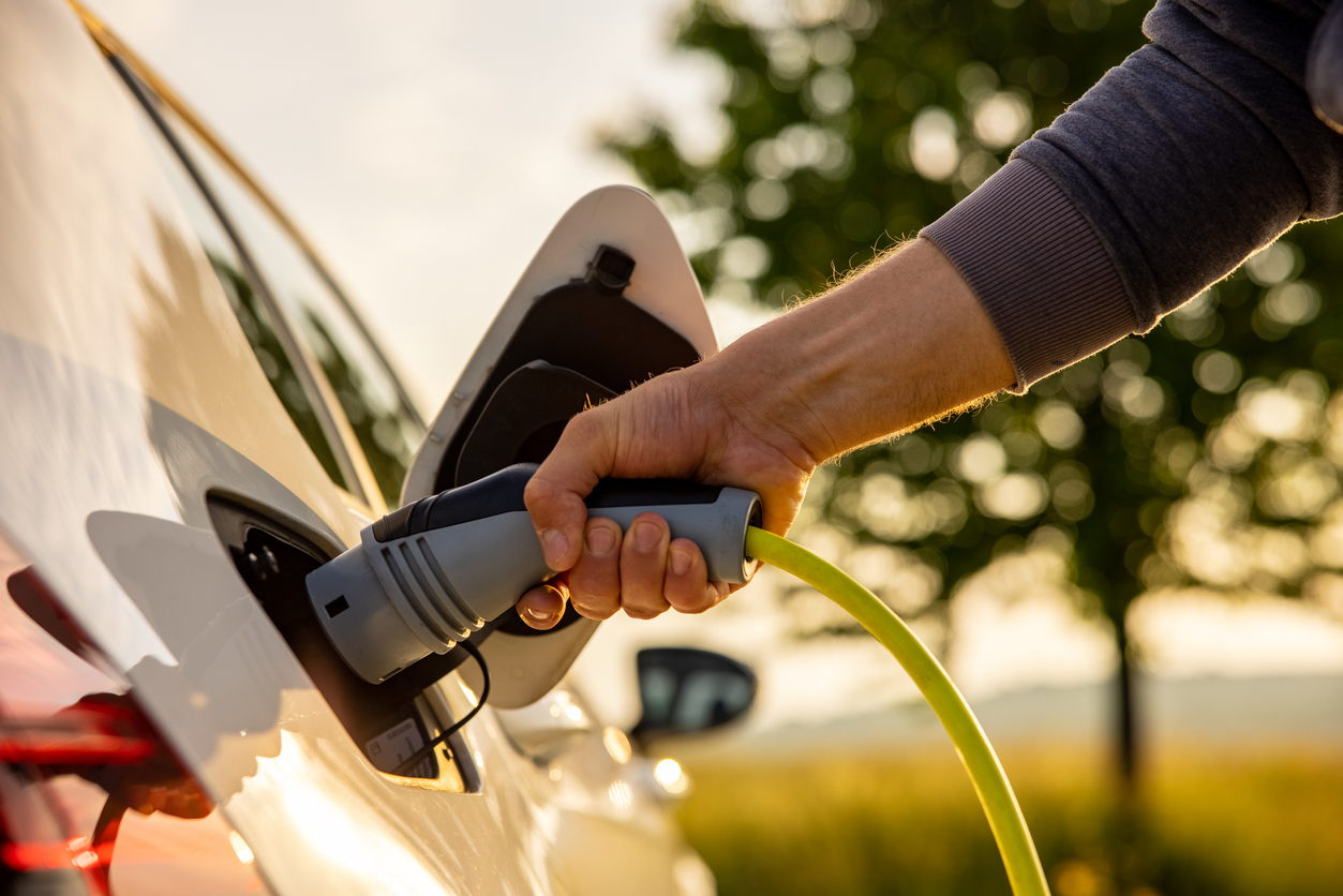 A close-up of a person’s hand plugging a yellow charging cable into an electric car at an outdoor charging station, with sunlight and greenery in the background.