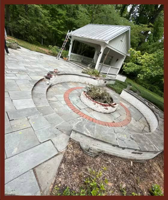 Backyard patio with large stone pavers and a curved built-in seating wall surrounding a circular brick fire pit, with a covered pergola structure and wooded landscape in the background.