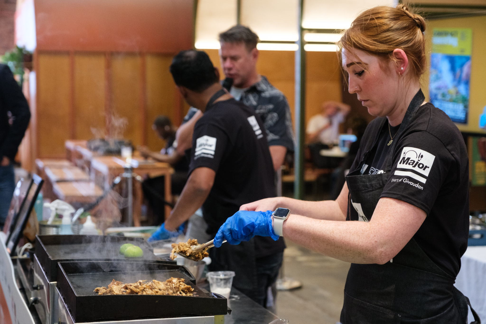 A woman wearing gloves and an apron cooks meat on a grill at an indoor food event, with other people and food stations in the background. Steam rises from the grill as she focuses on her task.