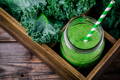 a green smoothie drink in a glass mason jar with a green and white striped straw on a wooden tray with leafy kale