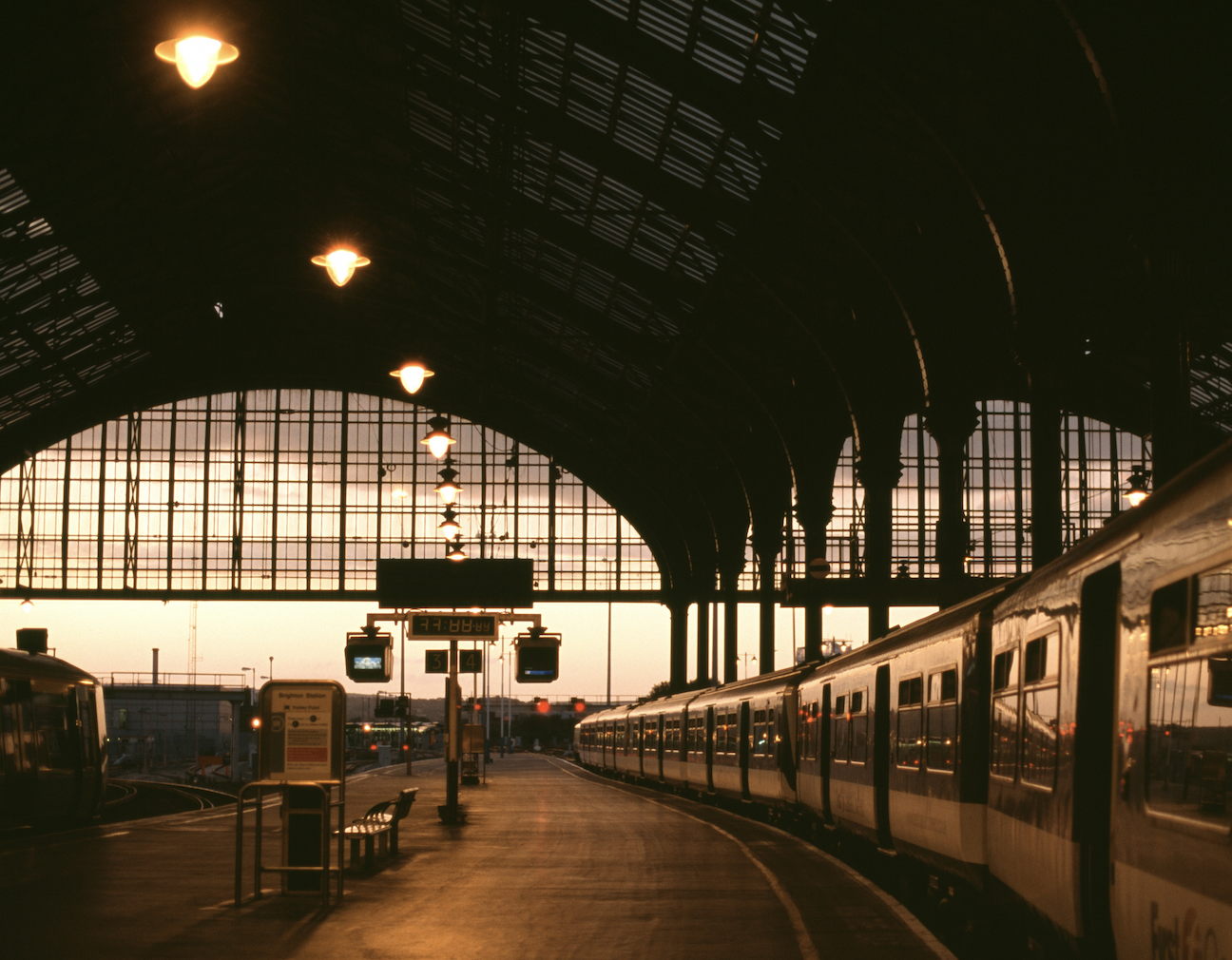 A train station at sunset with a curved glass roof, warmly lit lamps, and a train waiting on the platform. The scene is calm, with few people visible and soft, natural light streaming through the large windows.