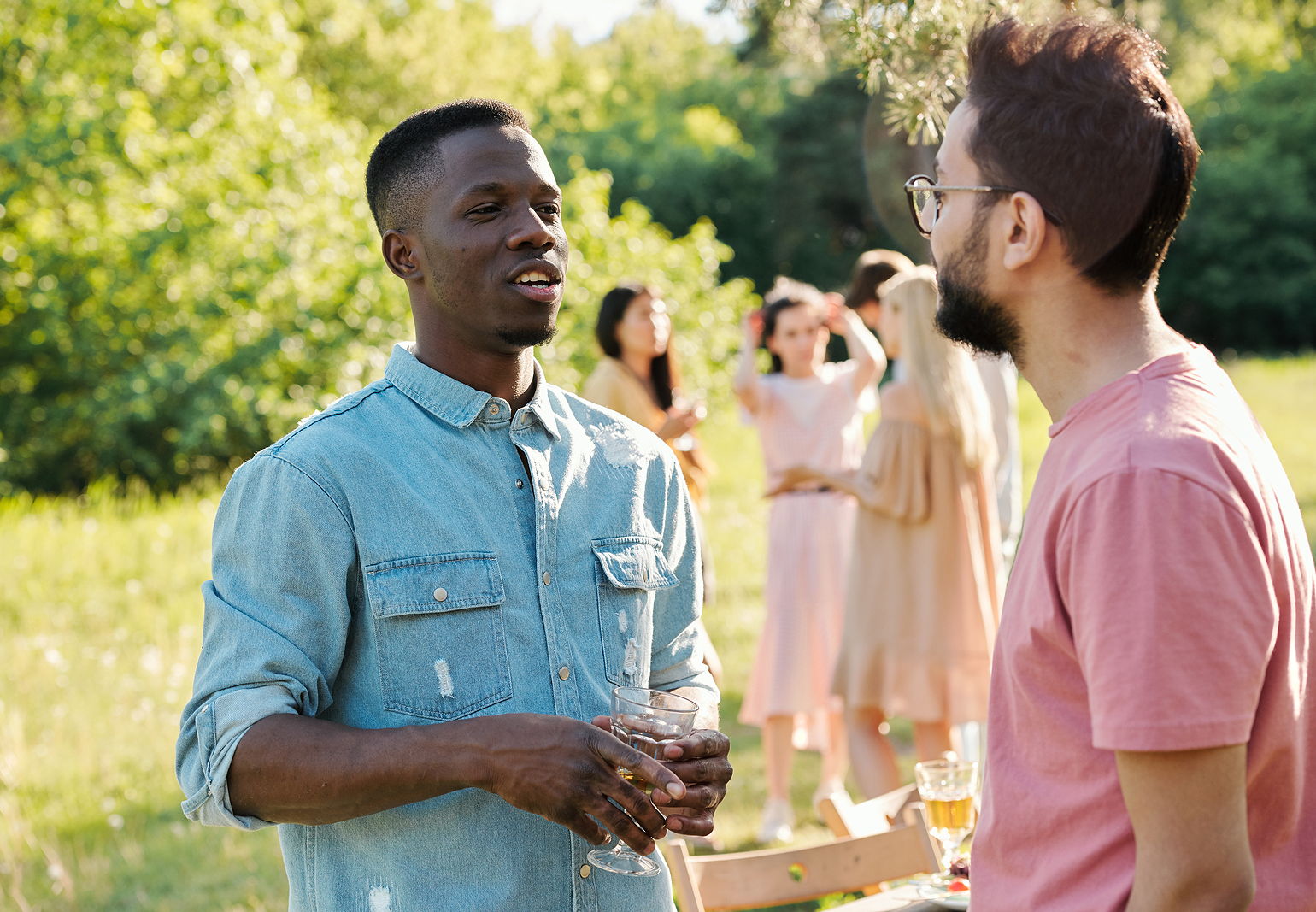 Photo of a young black and white man with glasses talking to each other at a friend reunion with girl friends in the background it a park.